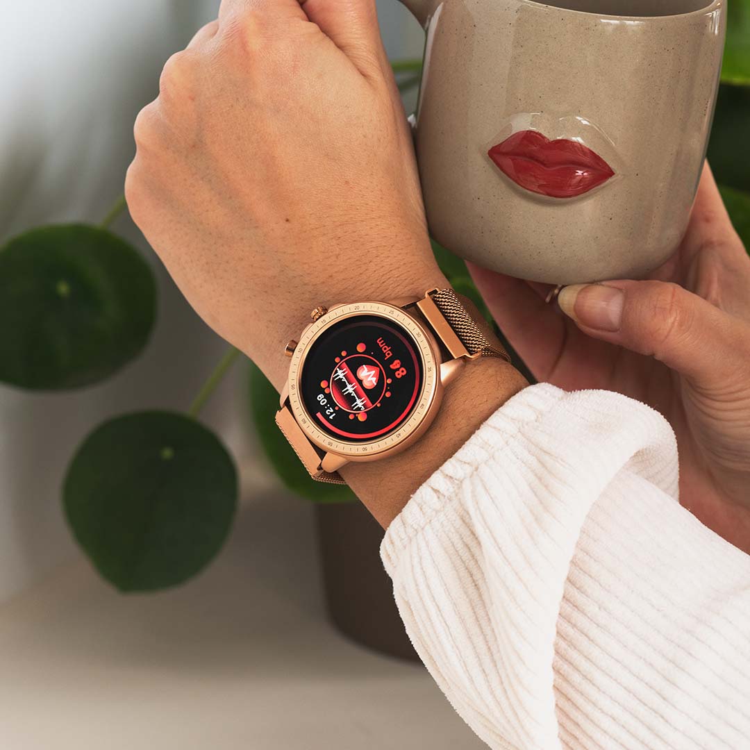 A person wearing a rose gold coloured OOZOO smartwatch with a metal mesh bracelet, displaying a colorful digital dial.